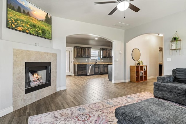 living room featuring a tiled fireplace, arched walkways, ceiling fan, and wood finished floors