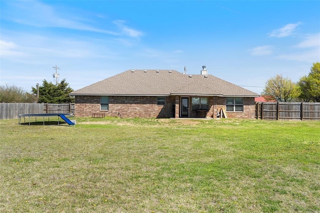 rear view of property with a yard, a fenced backyard, a trampoline, and roof with shingles