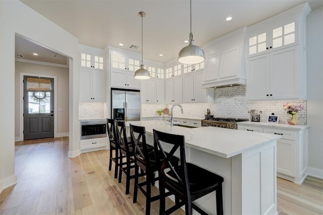 kitchen with a sink, an island with sink, stainless steel appliances, light wood-style flooring, and custom range hood