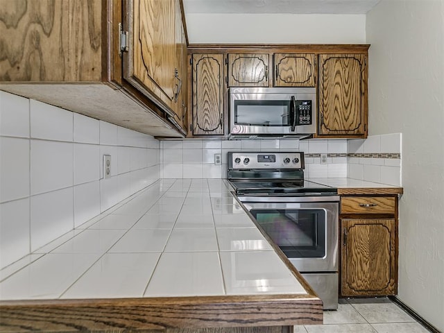 kitchen featuring tile countertops, light tile patterned floors, brown cabinetry, appliances with stainless steel finishes, and decorative backsplash