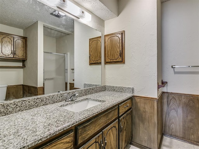 full bath with a textured wall, a textured ceiling, a shower stall, vanity, and a wainscoted wall
