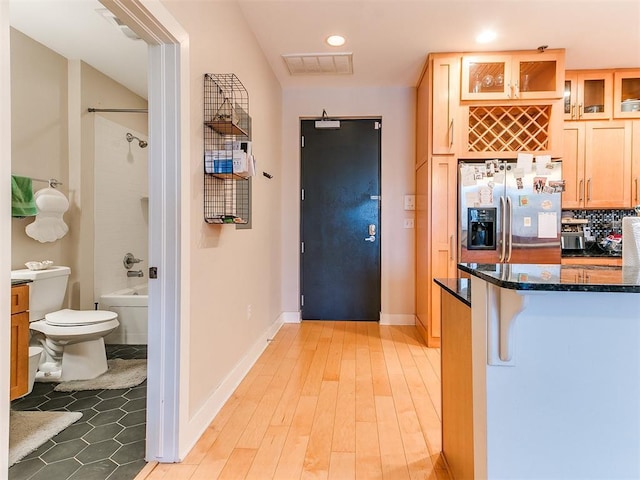 kitchen featuring dark stone counters, glass insert cabinets, light wood-type flooring, visible vents, and stainless steel refrigerator with ice dispenser