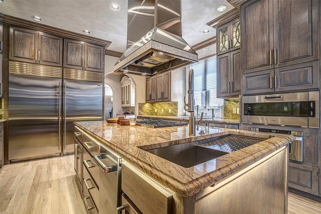5100 Wisteria Dr, Oklahoma City, OK 73142 - kitchen featuring light wood-type flooring, light stone counters, island range hood, a center island with sink, and stainless steel appliances