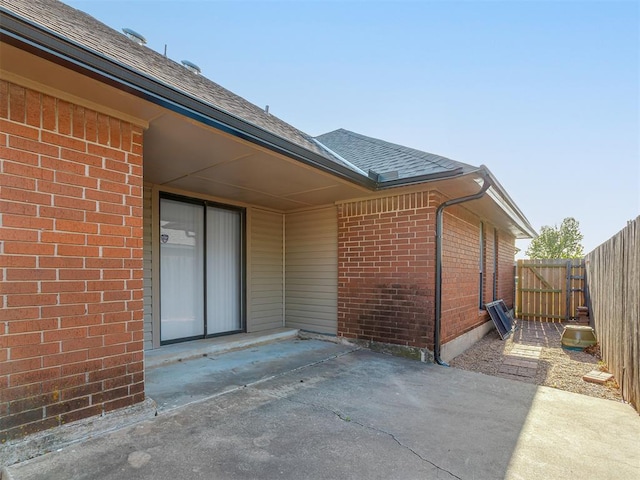 view of exterior entry featuring roof with shingles, a patio, brick siding, and fence