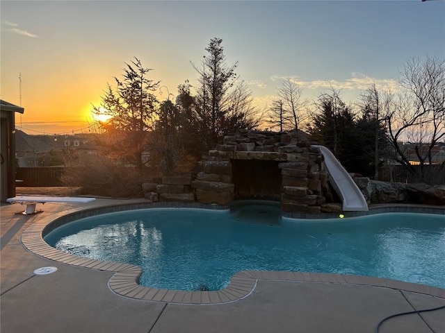 pool at dusk with a diving board, a water slide, and a fenced in pool