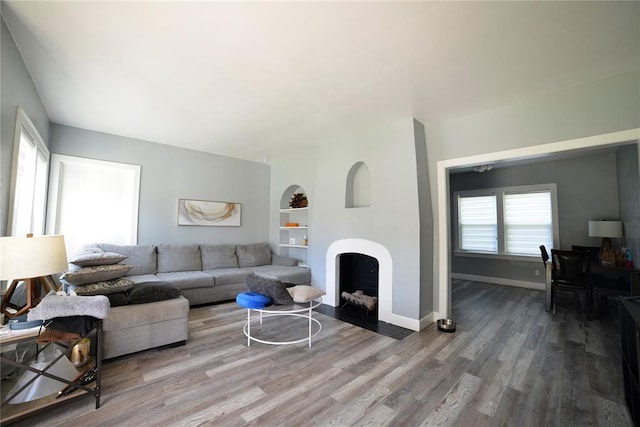 living room featuring a fireplace with flush hearth, built in shelves, baseboards, and wood finished floors