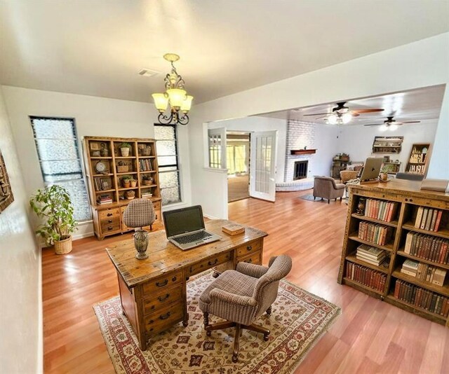 office area featuring light wood-type flooring, ceiling fan with notable chandelier, a brick fireplace, and visible vents