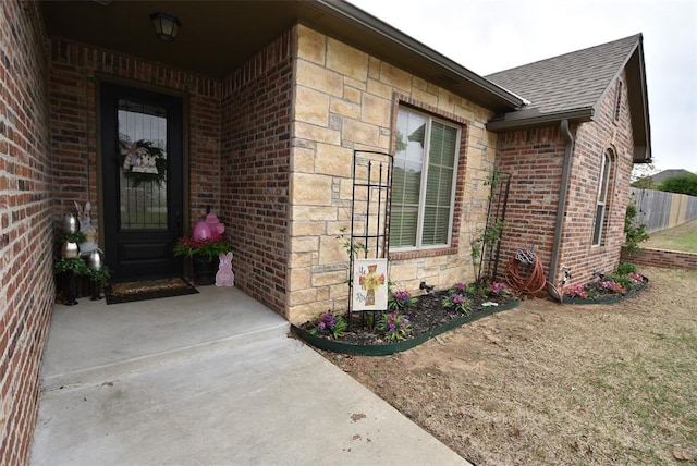 view of exterior entry featuring brick siding, fence, and a shingled roof