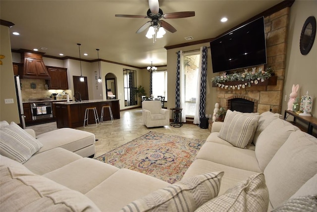 living area featuring a stone fireplace, a ceiling fan, crown molding, recessed lighting, and stone tile floors