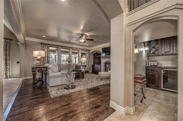 living room with a bar, ceiling fan, wine cooler, crown molding, and a glass covered fireplace