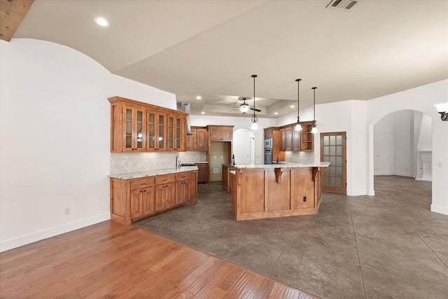 kitchen with a ceiling fan, brown cabinets, glass insert cabinets, arched walkways, and backsplash