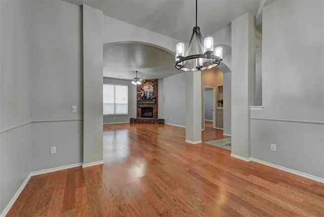 unfurnished dining area featuring wood finished floors, arched walkways, baseboards, a fireplace, and ceiling fan with notable chandelier