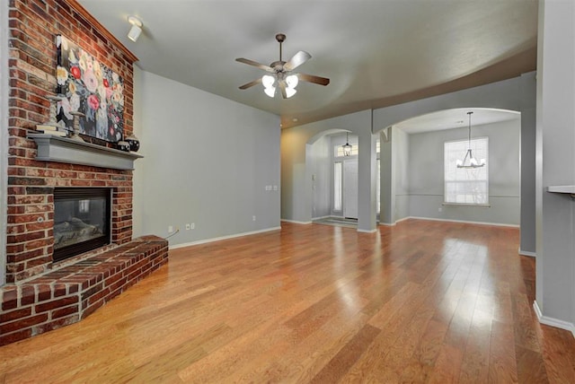 unfurnished living room with arched walkways, baseboards, ceiling fan with notable chandelier, wood finished floors, and a brick fireplace