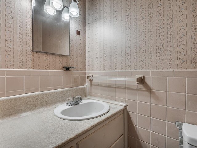 bathroom with tile walls, wallpapered walls, a wainscoted wall, and vanity