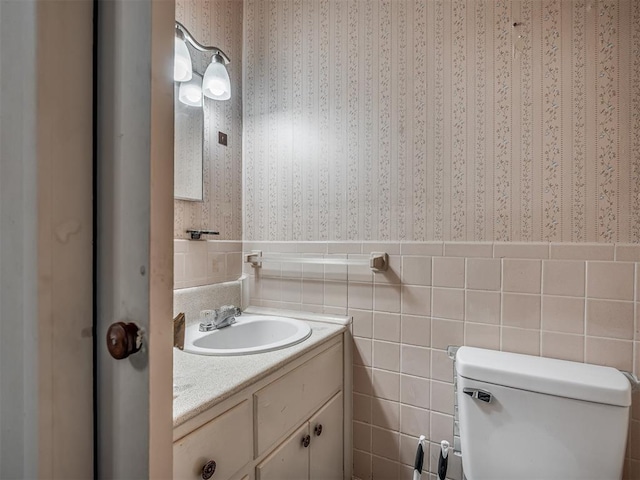 bathroom featuring wallpapered walls, vanity, tile walls, a wainscoted wall, and toilet