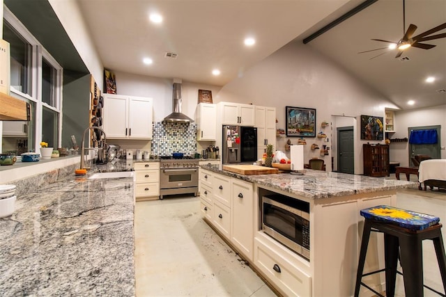 kitchen with a breakfast bar area, wall chimney exhaust hood, light stone counters, appliances with stainless steel finishes, and backsplash