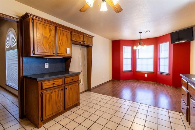 kitchen with dark countertops, ceiling fan with notable chandelier, brown cabinetry, light tile patterned flooring, and visible vents