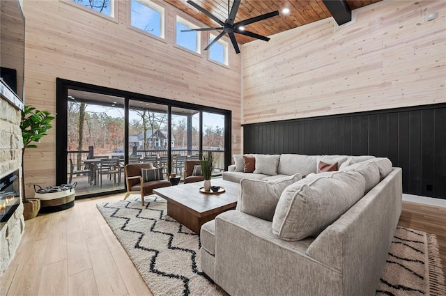 living room with a high ceiling, wood walls, a stone fireplace, a ceiling fan, and wooden ceiling