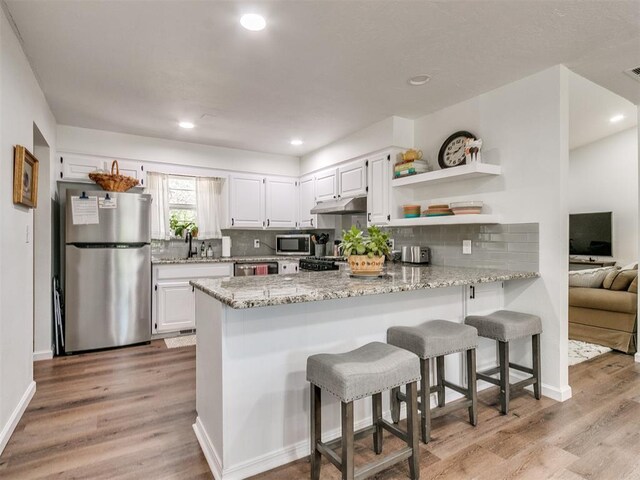 kitchen featuring open shelves, appliances with stainless steel finishes, light stone countertops, a peninsula, and under cabinet range hood