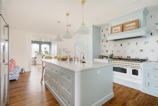 kitchen featuring wood finished floors, light stone countertops, an island with sink, range with two ovens, and hanging light fixtures