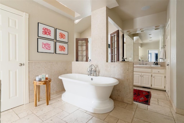 full bath with stone tile flooring, vanity, a freestanding bath, and tile walls