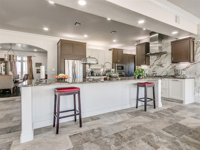 6363 NW 220th St, Edmond, OK 73025 - kitchen featuring wall chimney range hood, appliances with stainless steel finishes, a kitchen bar, backsplash, and ornamental molding