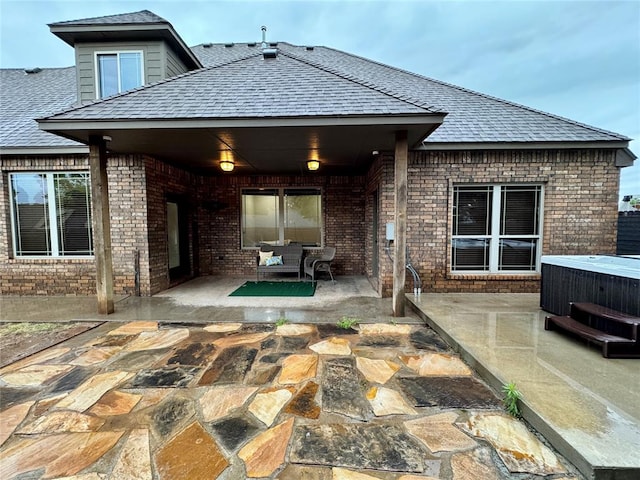 rear view of property with roof with shingles, a patio area, brick siding, and a hot tub