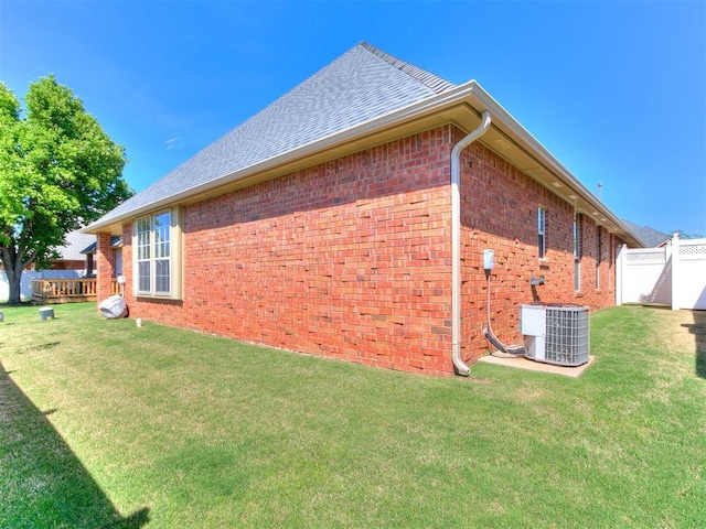 view of side of property with central AC, brick siding, fence, a yard, and roof with shingles
