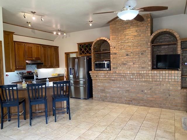kitchen with light tile patterned flooring, a kitchen bar, brick wall, appliances with stainless steel finishes, and ceiling fan