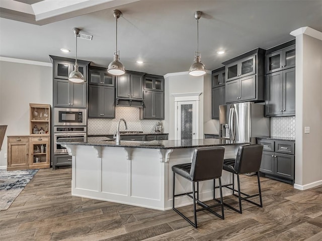 kitchen with stainless steel appliances, dark stone countertops, a sink, a center island with sink, and tasteful backsplash