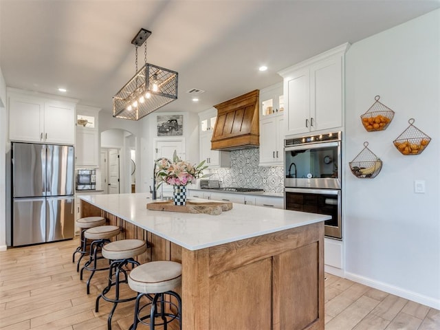 kitchen featuring premium range hood, arched walkways, light wood-type flooring, decorative backsplash, and appliances with stainless steel finishes