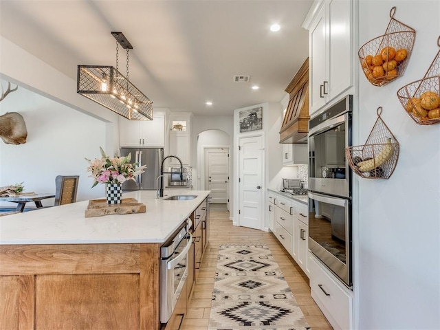 kitchen with a sink, a kitchen island with sink, arched walkways, stainless steel appliances, and light wood-style flooring