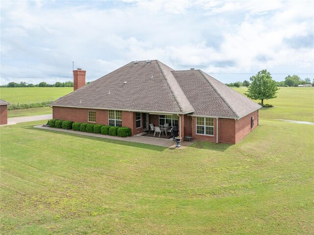 back of house featuring roof with shingles, a chimney, brick siding, a patio area, and a yard