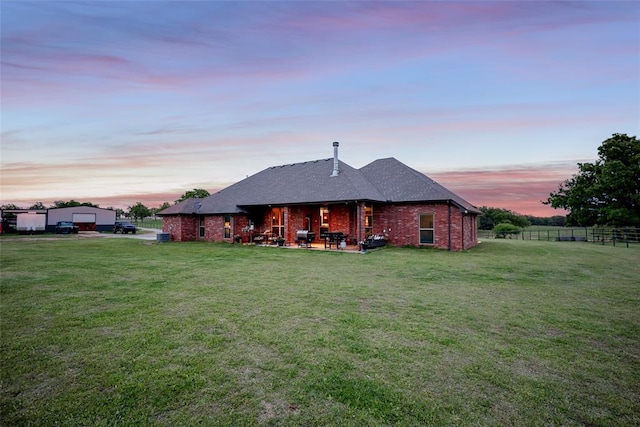 back of property at dusk featuring roof with shingles, a lawn, brick siding, fence, and a patio