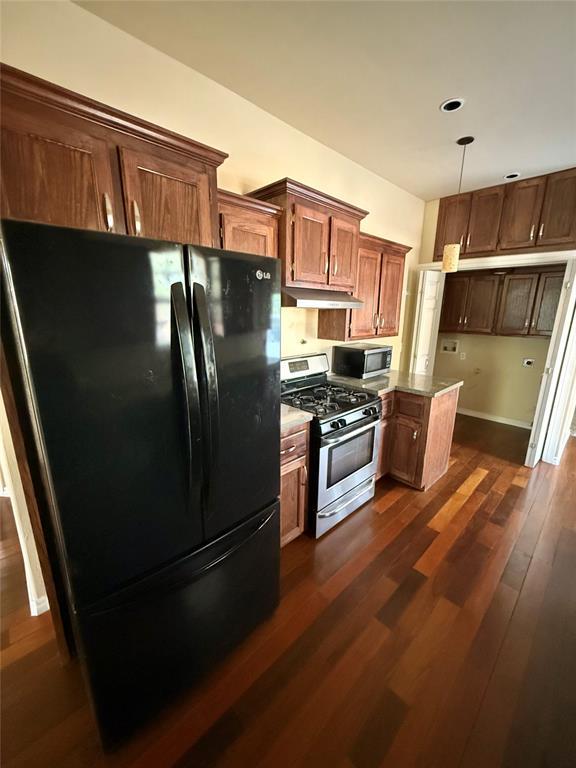 kitchen with brown cabinets, dark wood-style floors, under cabinet range hood, stainless steel appliances, and hanging light fixtures