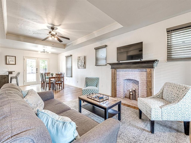 living room with a brick fireplace, a tray ceiling, crown molding, a ceiling fan, and wood finished floors