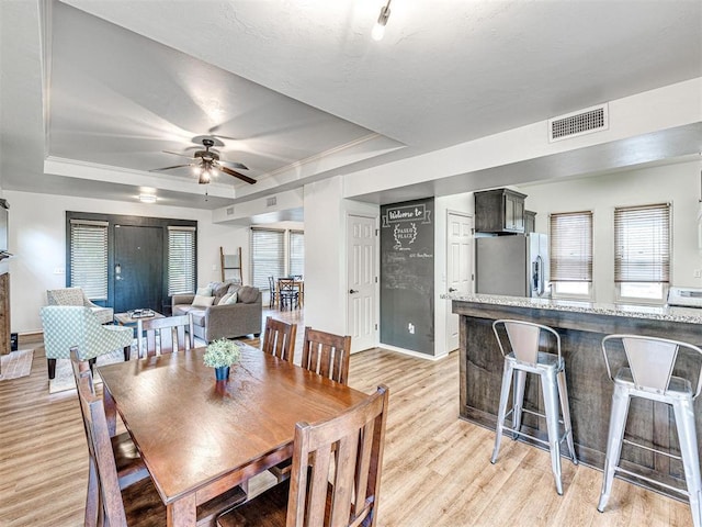 dining space featuring visible vents, a tray ceiling, baseboards, light wood-type flooring, and a ceiling fan
