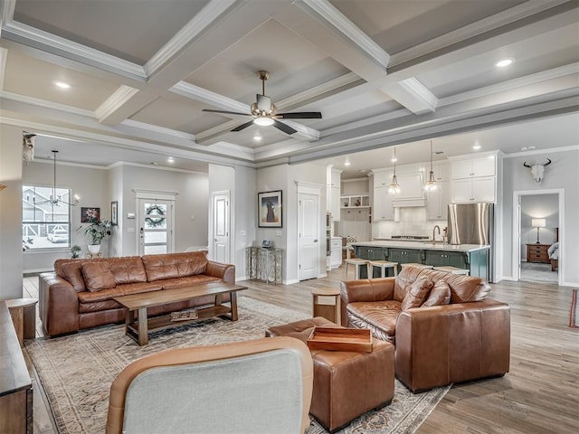 living area with beam ceiling, light wood-style floors, ceiling fan with notable chandelier, ornamental molding, and coffered ceiling