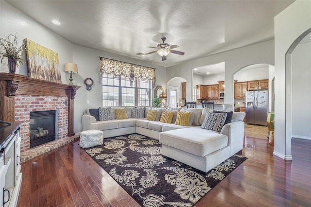 living area featuring hardwood / wood-style floors, a ceiling fan, arched walkways, baseboards, and a brick fireplace