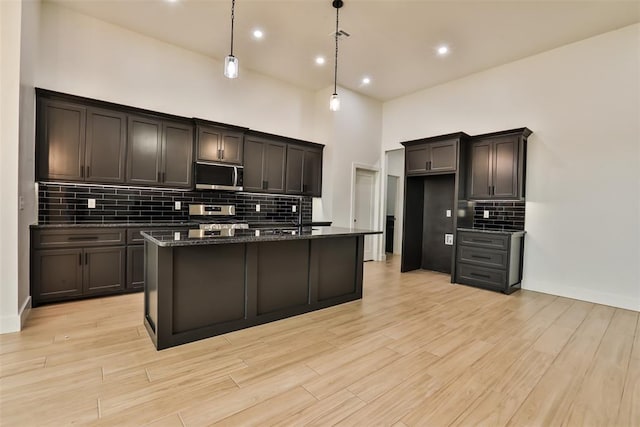 kitchen featuring light wood-style floors, a center island with sink, tasteful backsplash, dark stone countertops, and appliances with stainless steel finishes