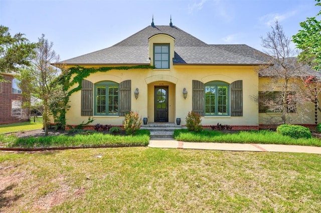 french country style house featuring a shingled roof, a front lawn, and stucco siding