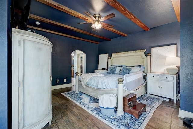 bedroom with wood finished floors, arched walkways, a textured wall, and beam ceiling