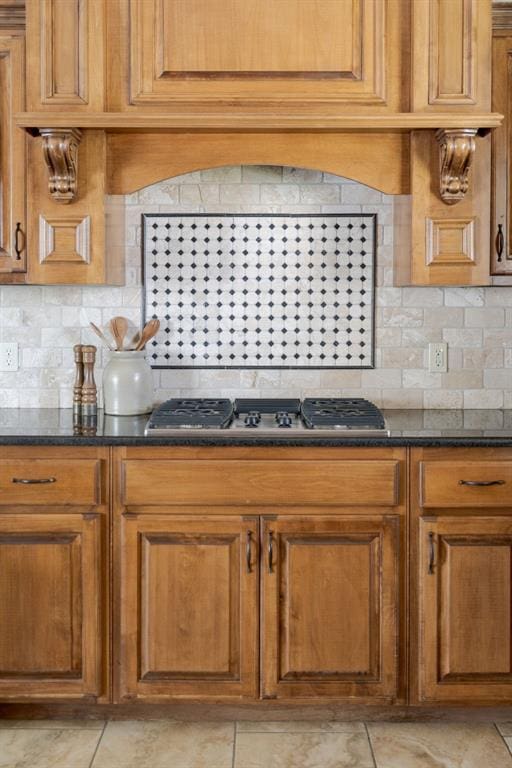 kitchen featuring brown cabinetry, backsplash, stainless steel gas stovetop, and dark stone counters