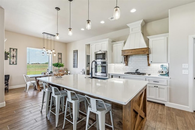 kitchen featuring a sink, a kitchen island with sink, baseboards, custom range hood, and appliances with stainless steel finishes