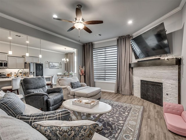 living area with visible vents, ceiling fan with notable chandelier, a stone fireplace, light wood-type flooring, and crown molding