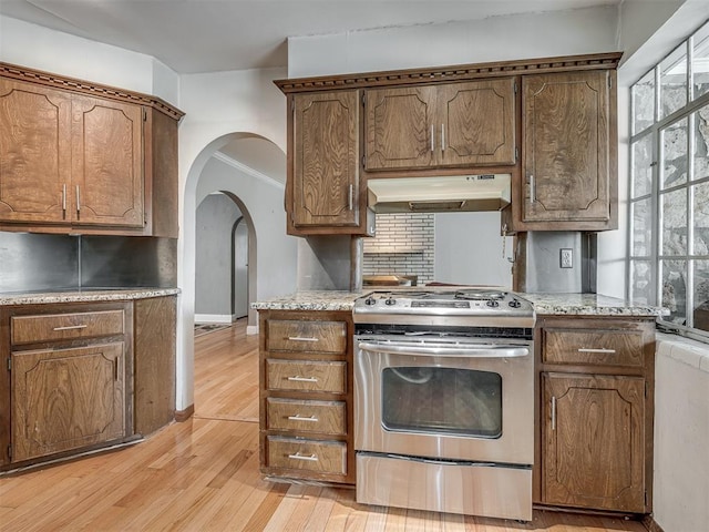 kitchen with exhaust hood, light wood-type flooring, stainless steel gas stove, light countertops, and arched walkways