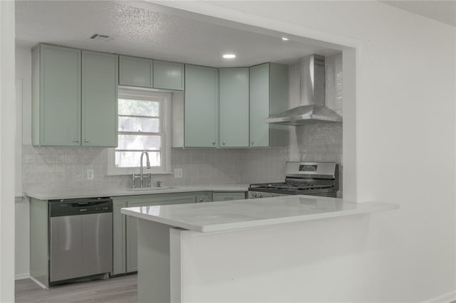 kitchen with stainless steel appliances, light countertops, wall chimney range hood, a sink, and a peninsula