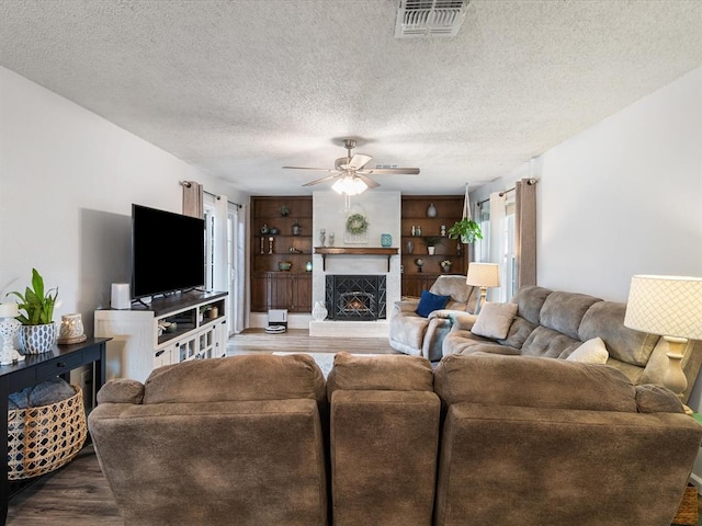 living area featuring ceiling fan, a textured ceiling, visible vents, wood finished floors, and a warm lit fireplace