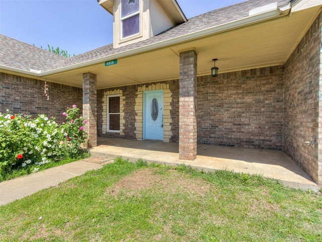 view of exterior entry with roof with shingles, covered porch, and brick siding