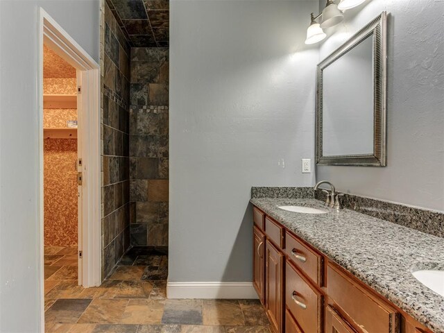 bathroom with a sink, stone finish floor, a tile shower, and double vanity